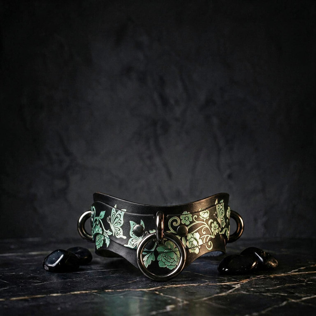 A black leather choker necklace with a floral design and two circular metal rings, placed on a dark surface with small black stones or crystals around it.