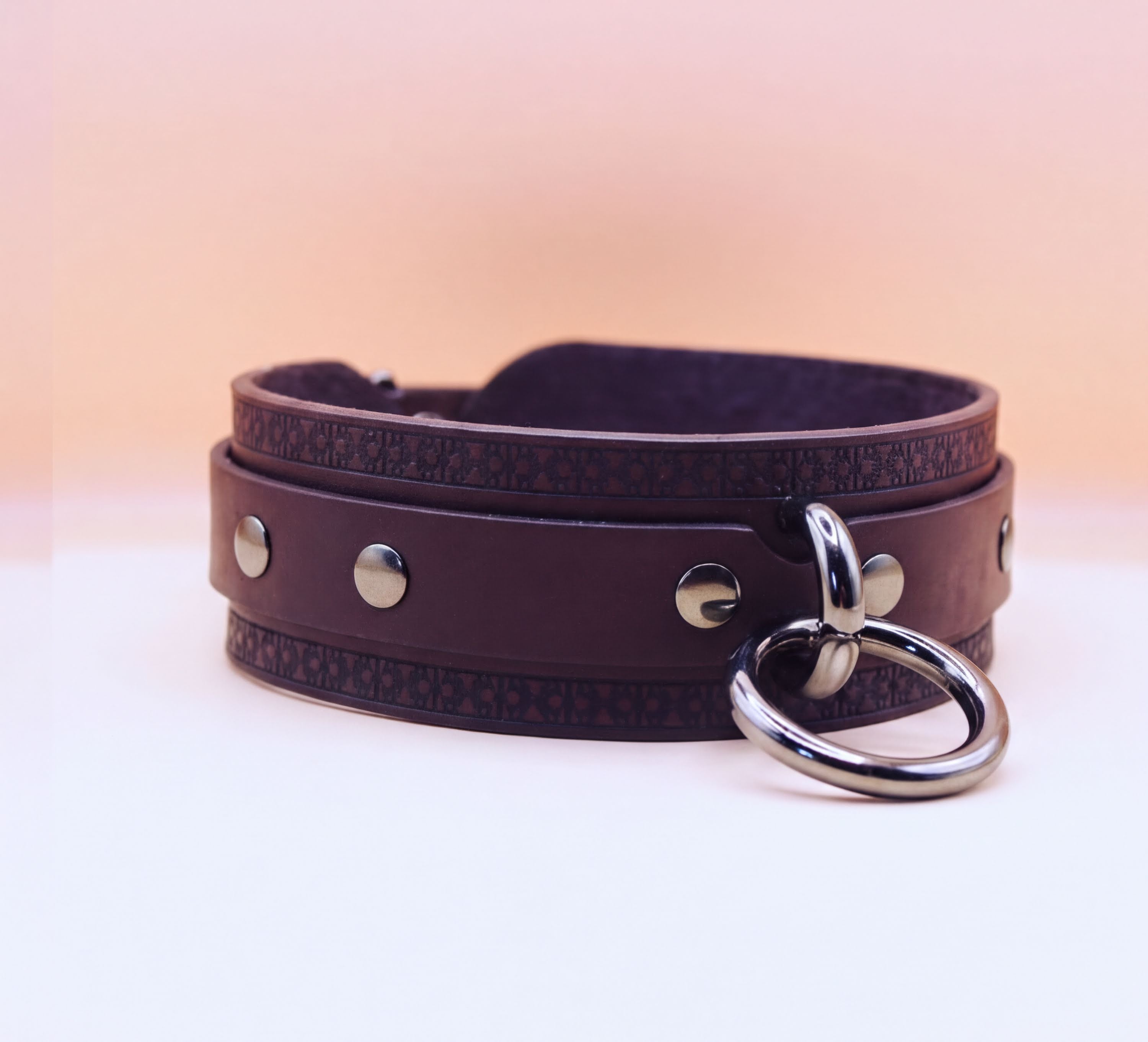 A brown leather choker collar with metal studs and a metal ring hanging from it.