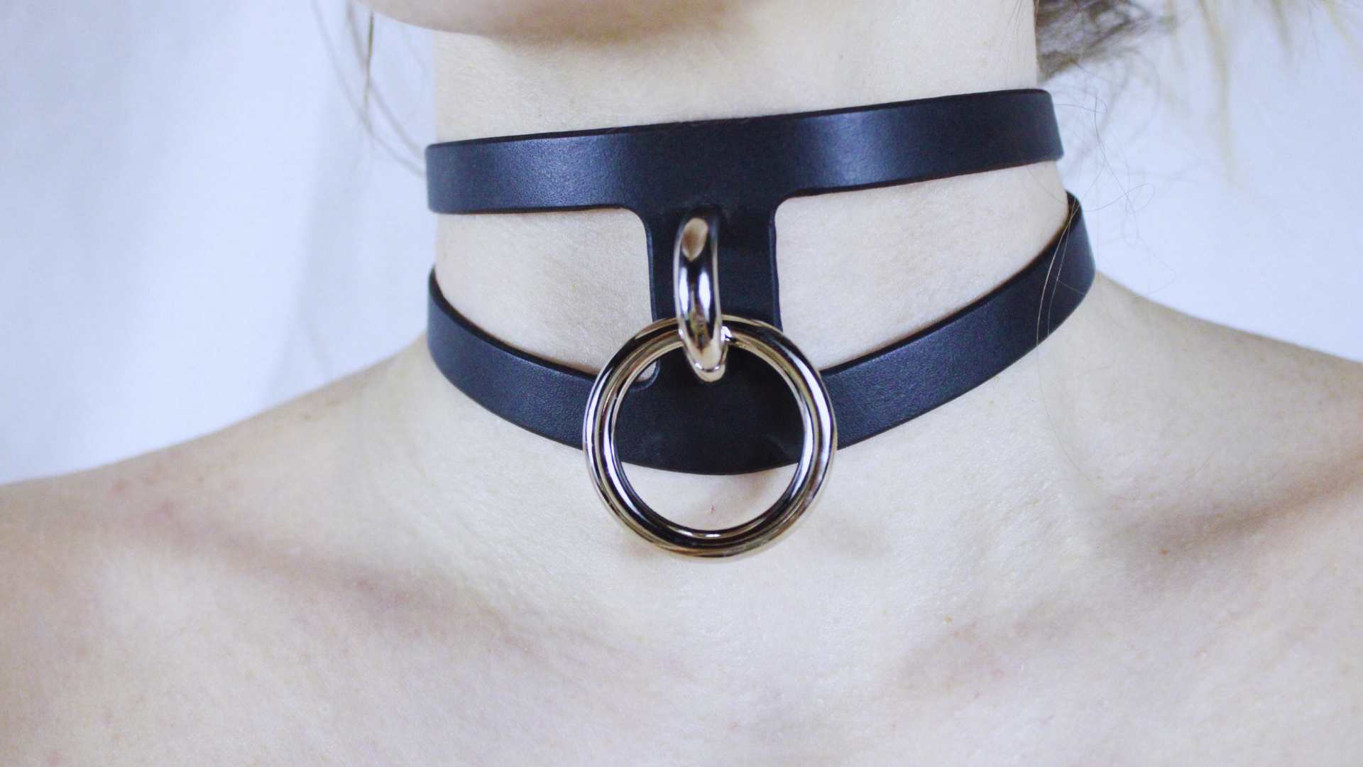 A close-up view of a person's neck and chest area wearing a black leather choker necklace with a large metal ring.