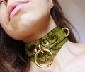 A close-up of a woman's neck and chin wearing a green choker necklace with a skull design and gold-colored metal rings.