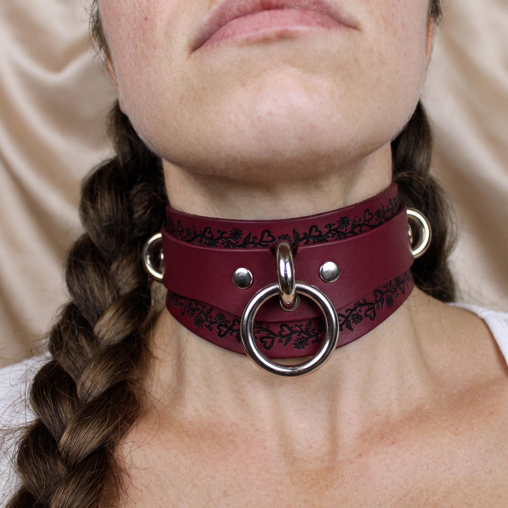 A close-up of a woman's neck wearing a burgundy leather choker necklace with a metal ring and two metal rings on either side.