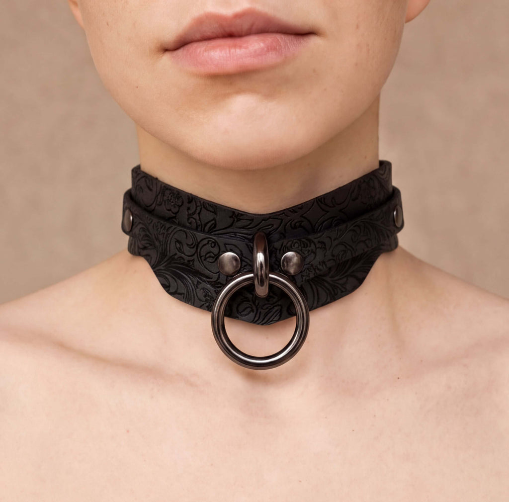 A close-up of a person's neck and chest area wearing a black choker necklace with a metal ring.