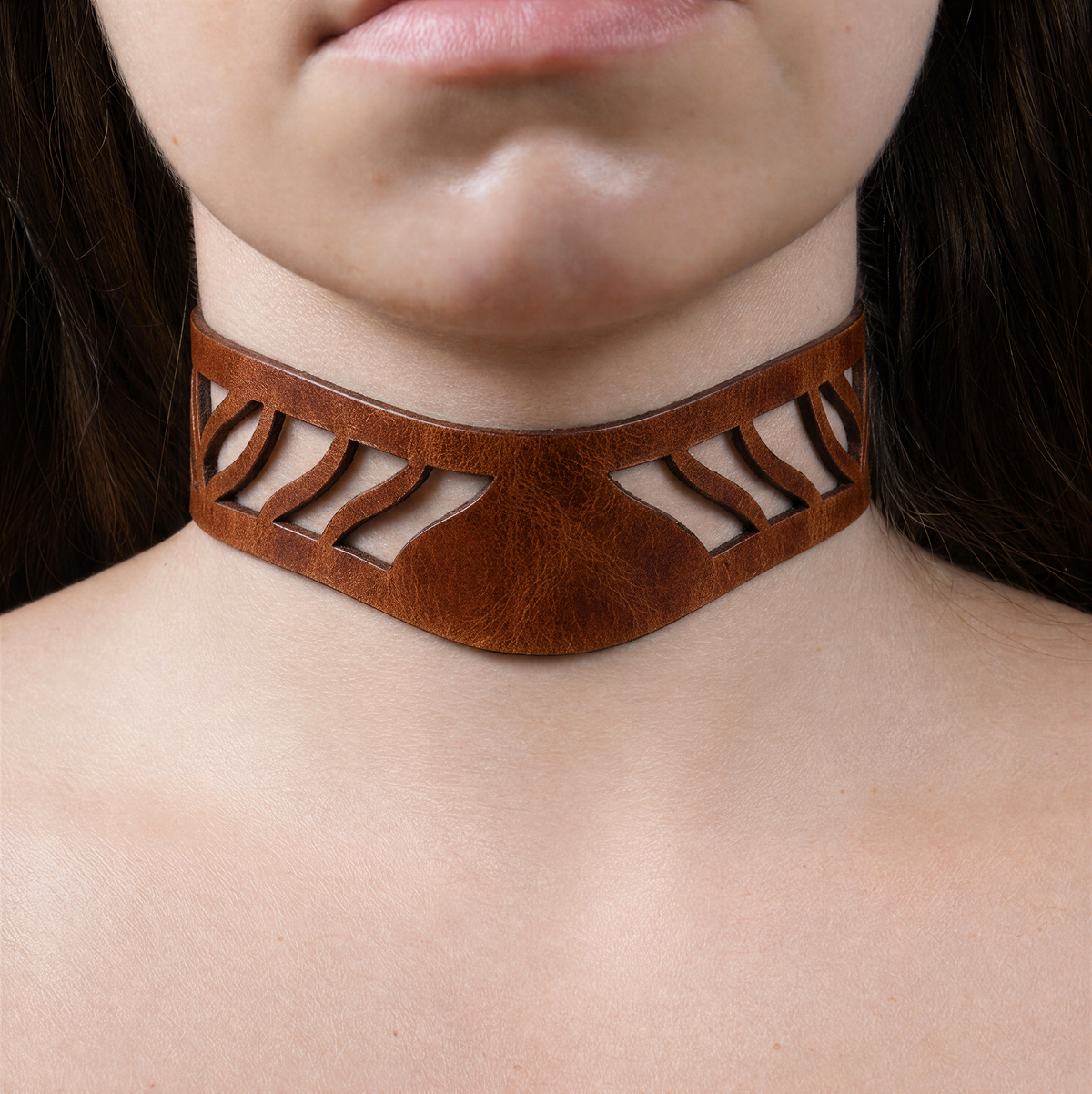 A close-up of a person's neck and chest area wearing a brown leather choker necklace with a unique cutout design.