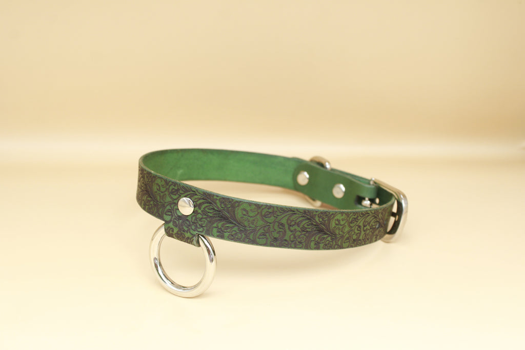 A green leather collar with a metal ring and buckle, placed on a beige background.