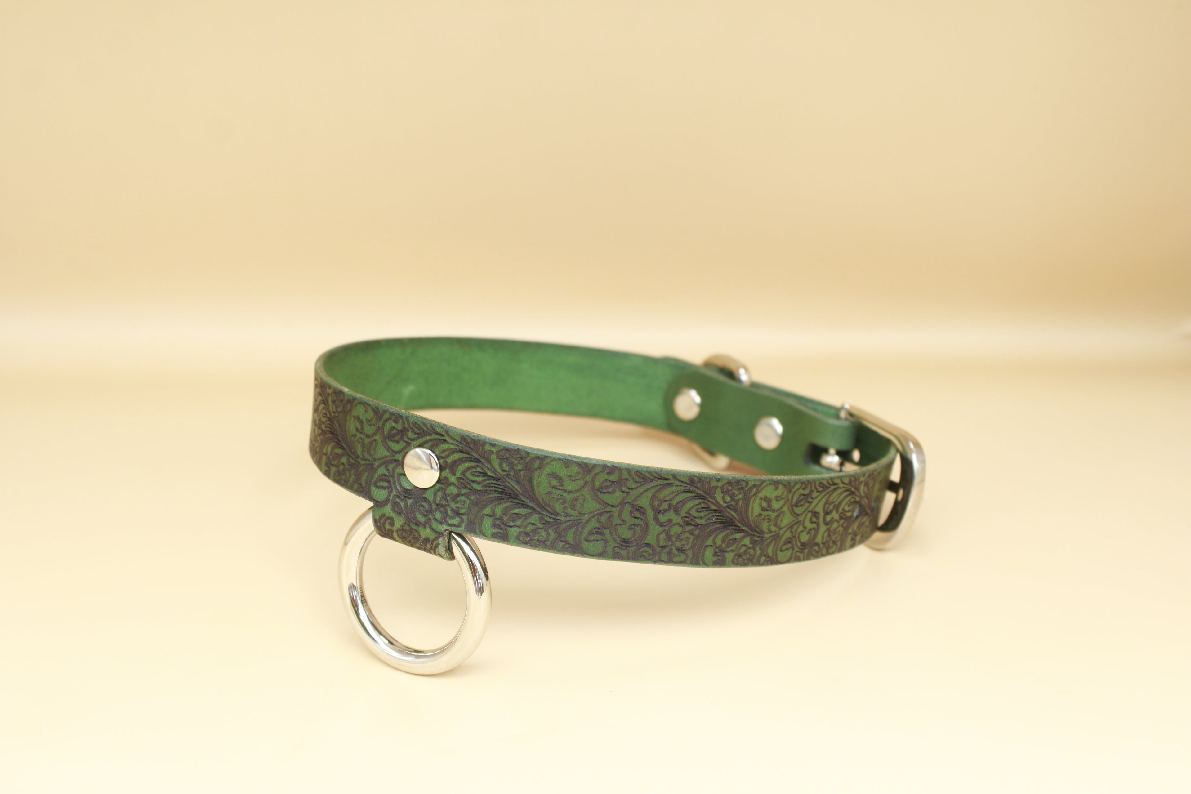 A green leather collar with a metal ring and buckle, placed on a beige background.