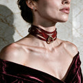 A woman wearing a red choker necklace with a gold buckle, posing against a marble wall.