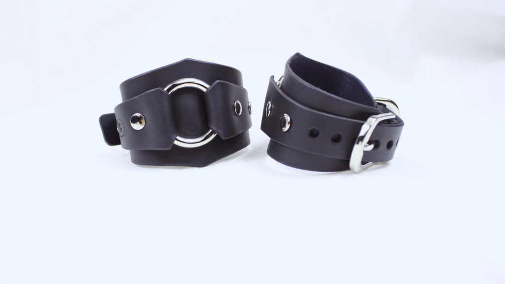A pair of black leather cuffs with metal rings and buckles, placed side by side on a white background.
