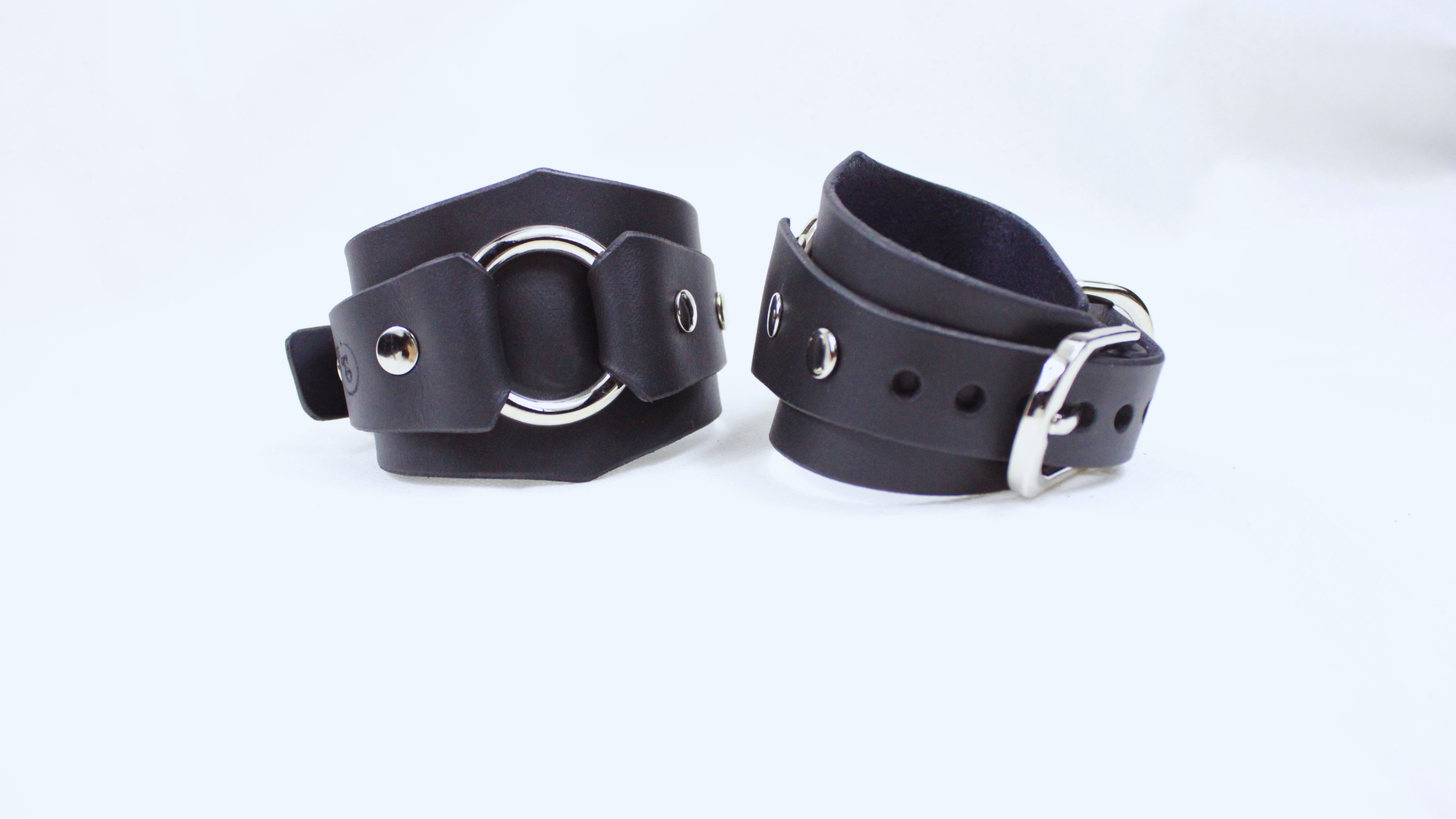 A pair of black leather cuffs with metal rings and buckles, placed side by side on a white background.