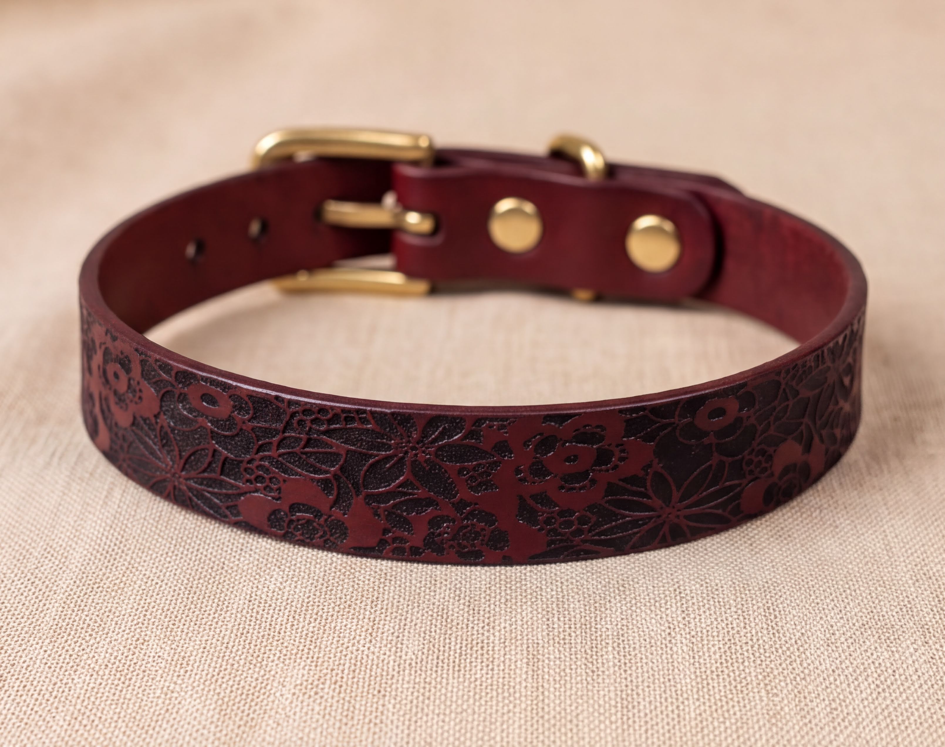 A burgundy leather dog collar with a floral pattern and a gold buckle is displayed on a beige fabric background.