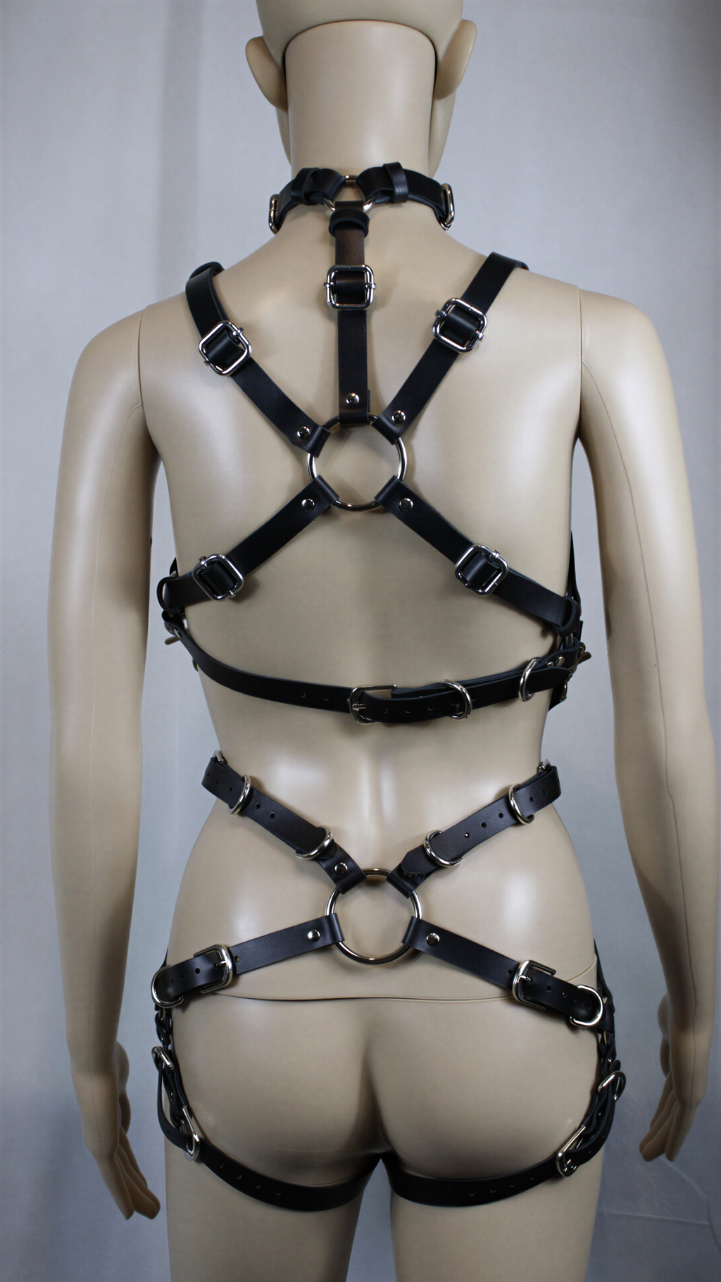A mannequin wearing a leather harness with multiple straps and buckles, including a cross-shaped strap across the back.