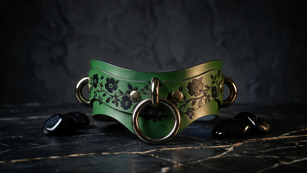 A green leather collar with floral designs and metal rings is displayed on a dark surface, accompanied by black stones.