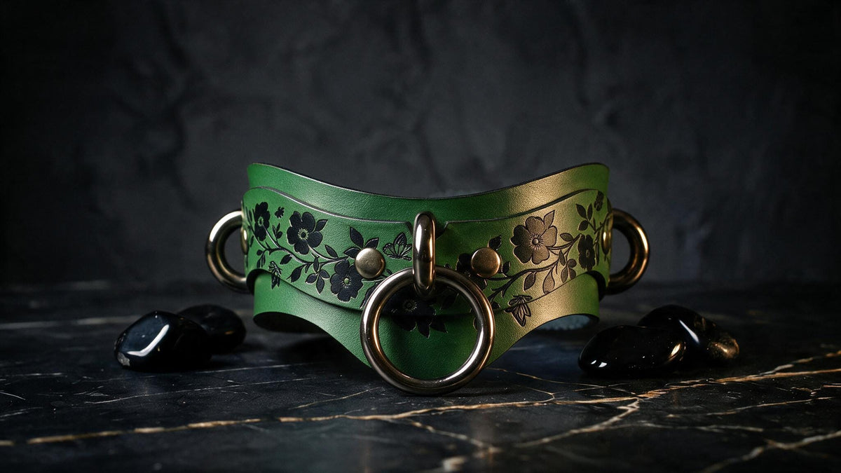 A green leather collar with floral designs and metal rings is displayed on a dark surface, accompanied by black stones.