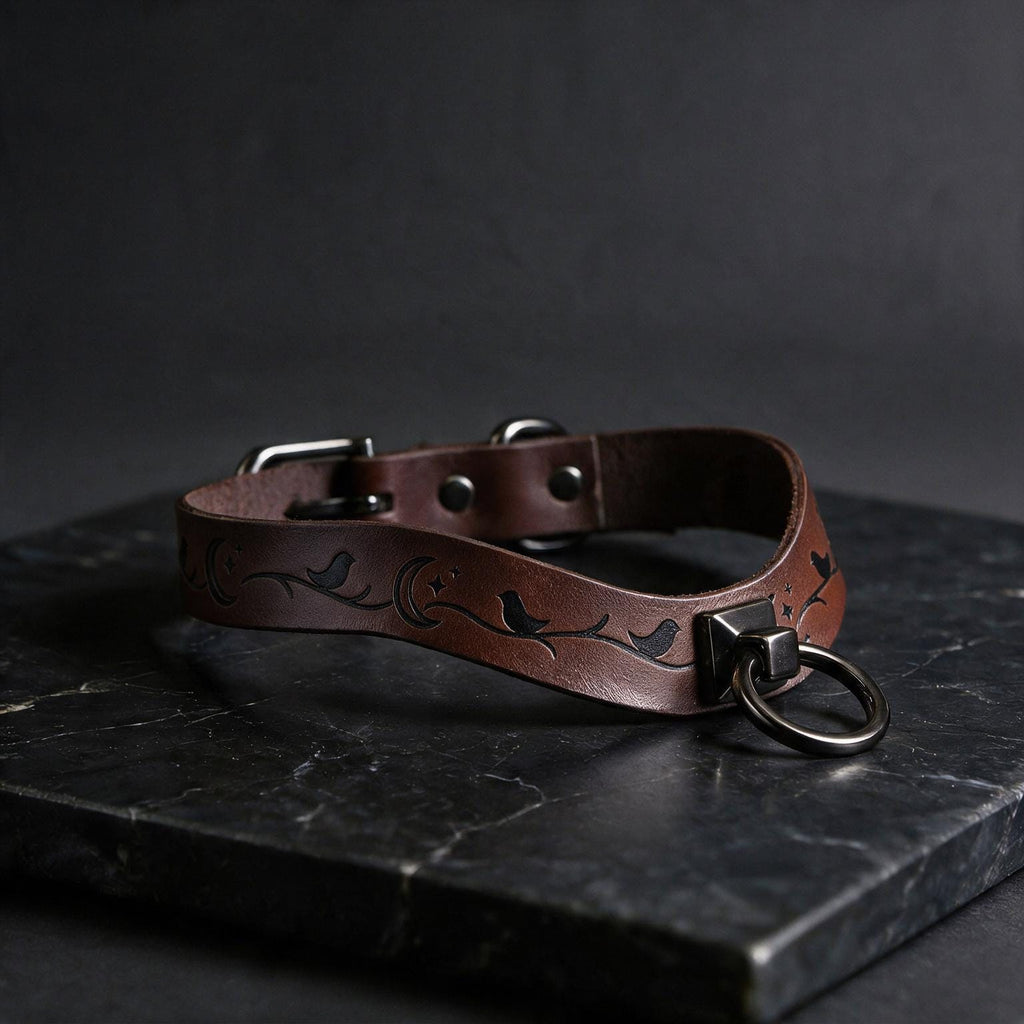 A brown leather collar with a metal ring and a bird design, resting on a dark surface.