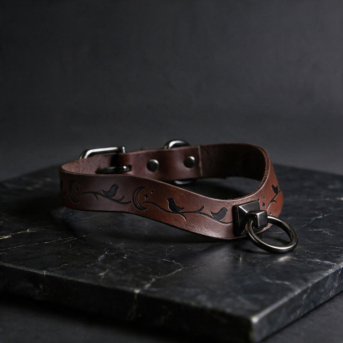 A brown leather collar with a metal ring and a bird design, resting on a dark surface.