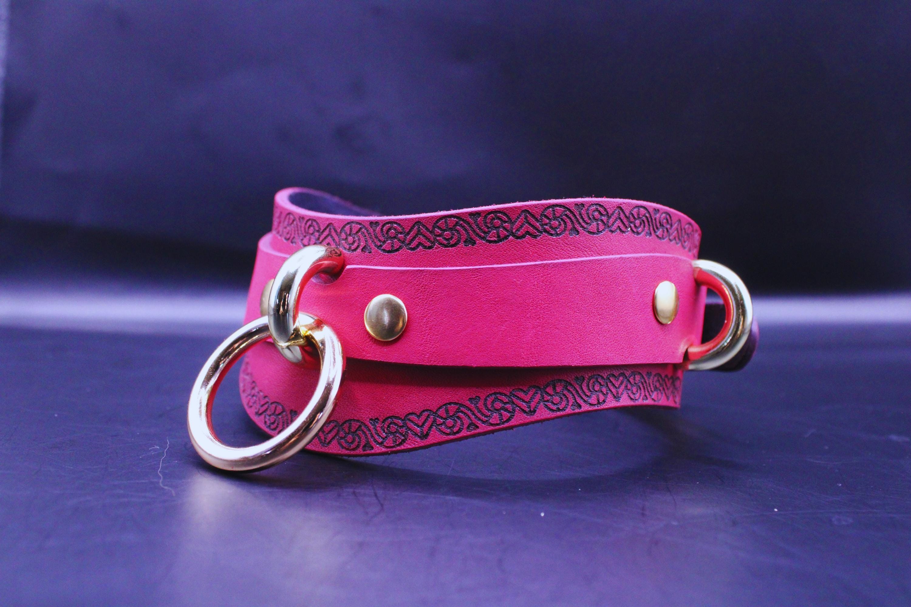 A pink leather choker necklace with a decorative pattern around the edges and two gold-colored rings on the front.