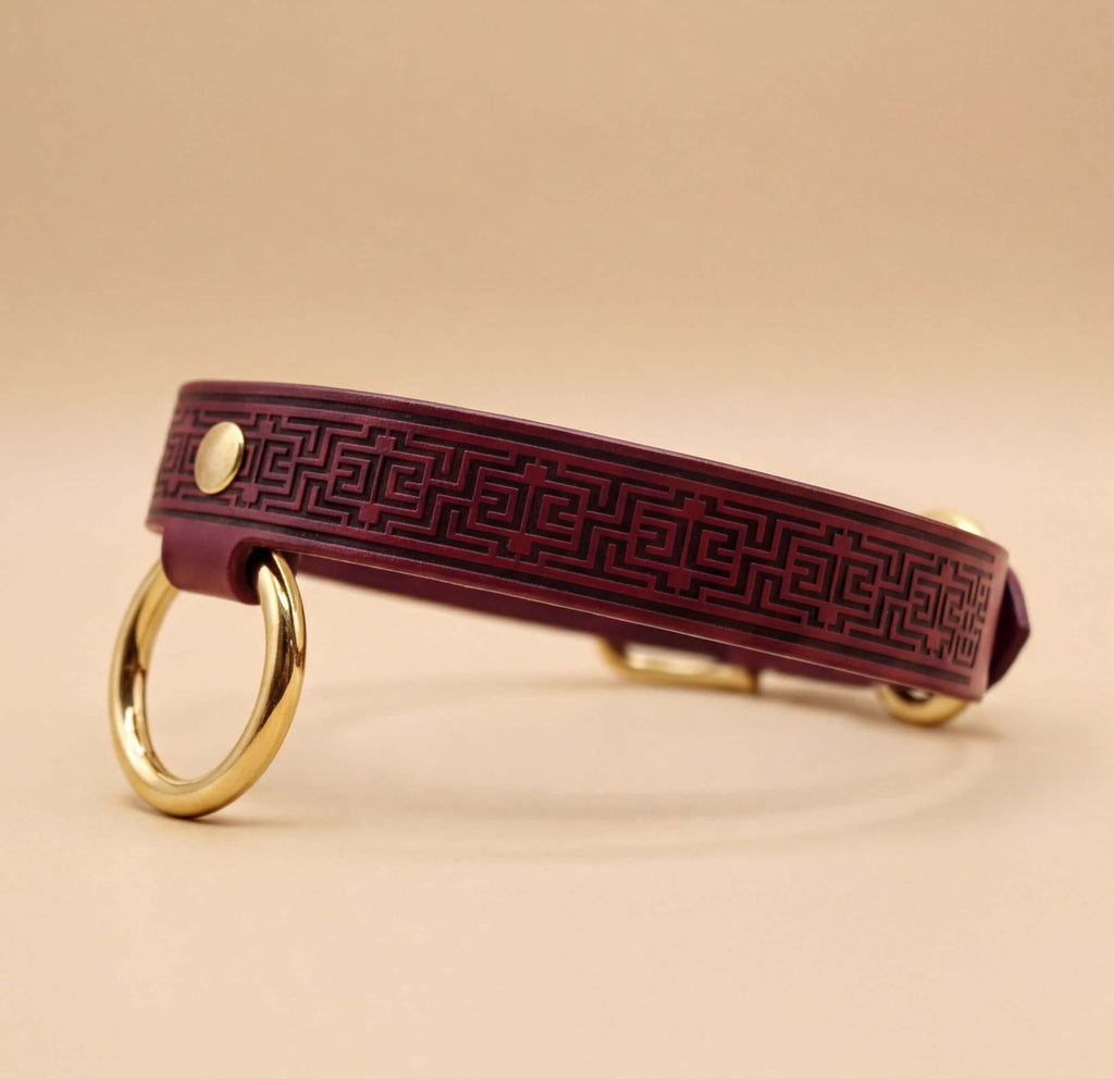 A burgundy leather dog collar with a geometric pattern and two gold-colored rings on either side.
