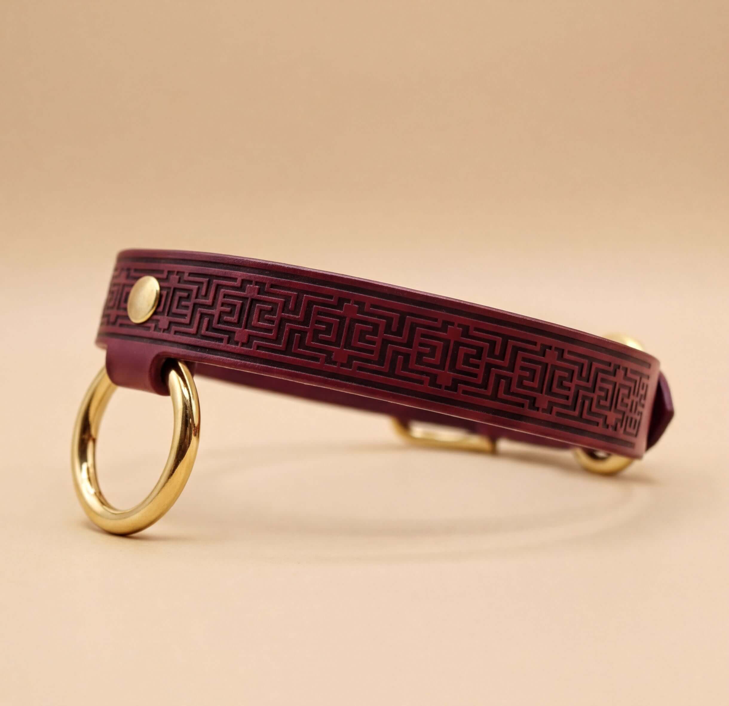 A burgundy leather dog collar with a geometric pattern and two gold-colored rings on either side.