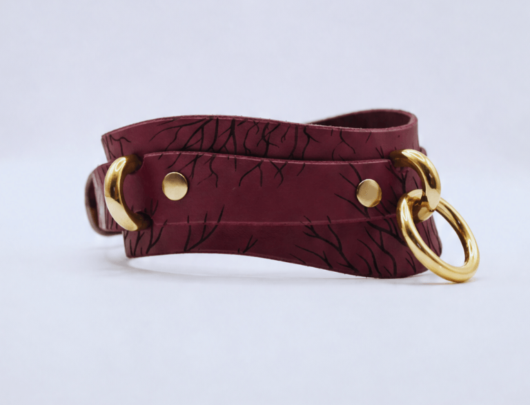 A burgundy leather choker necklace with a gold-colored metal ring and two gold-colored metal rings on either side.