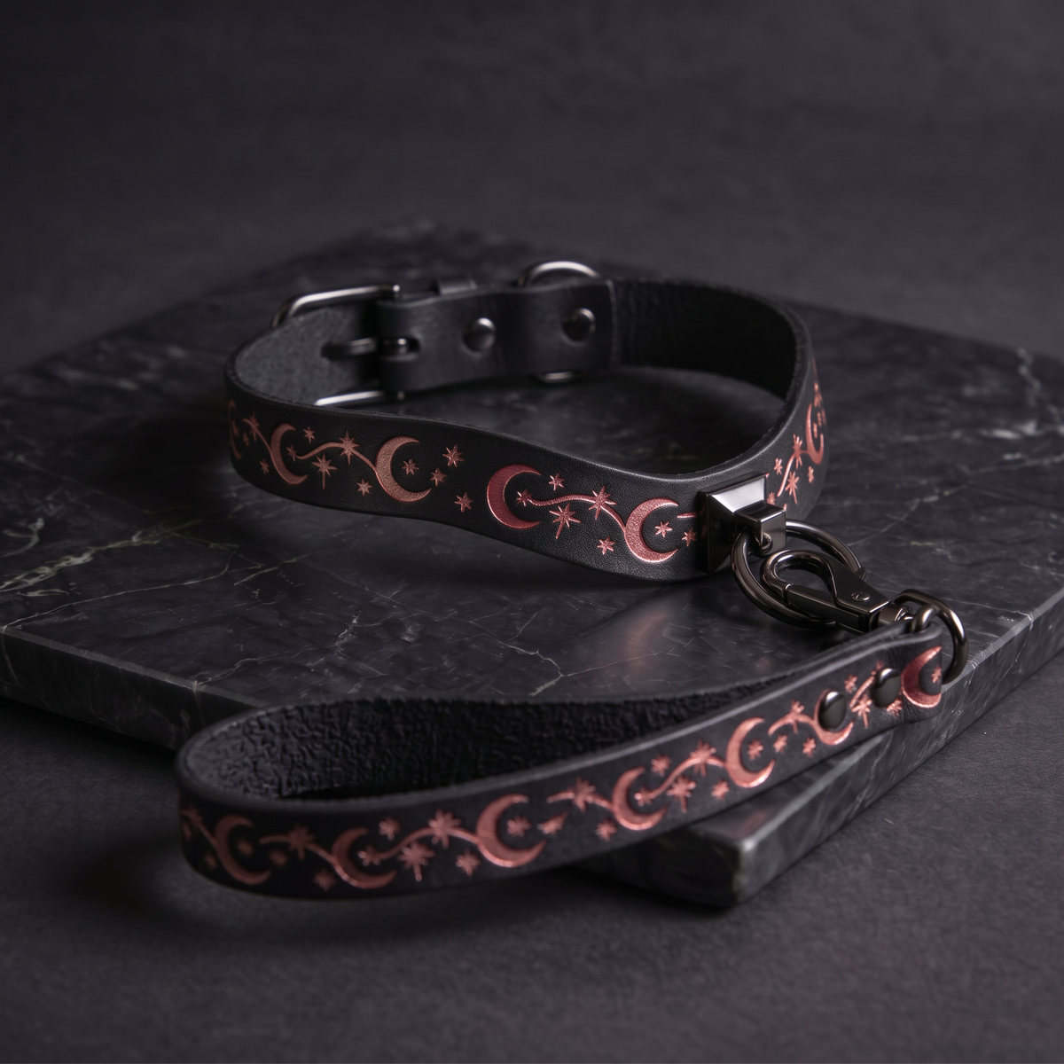 A black leather collar with a decorative pattern of stars and crescent moons, and a matching leash, resting on a dark surface.