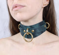 A close-up of a woman's neck and chest area wearing a dark green choker necklace with gold-colored metal rings.