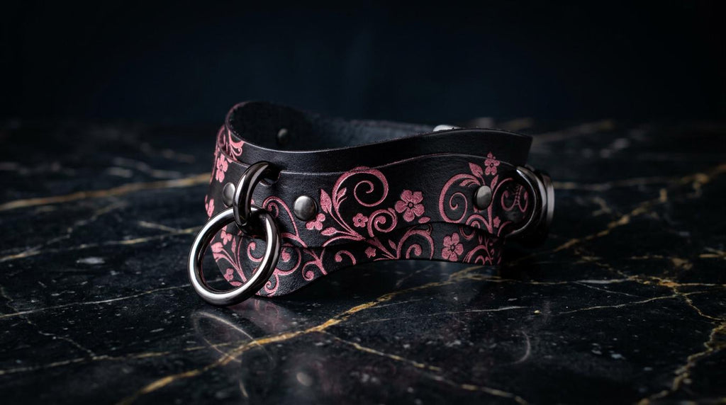 A black leather choker necklace with a floral pattern and metal rings is displayed on a black marble surface.