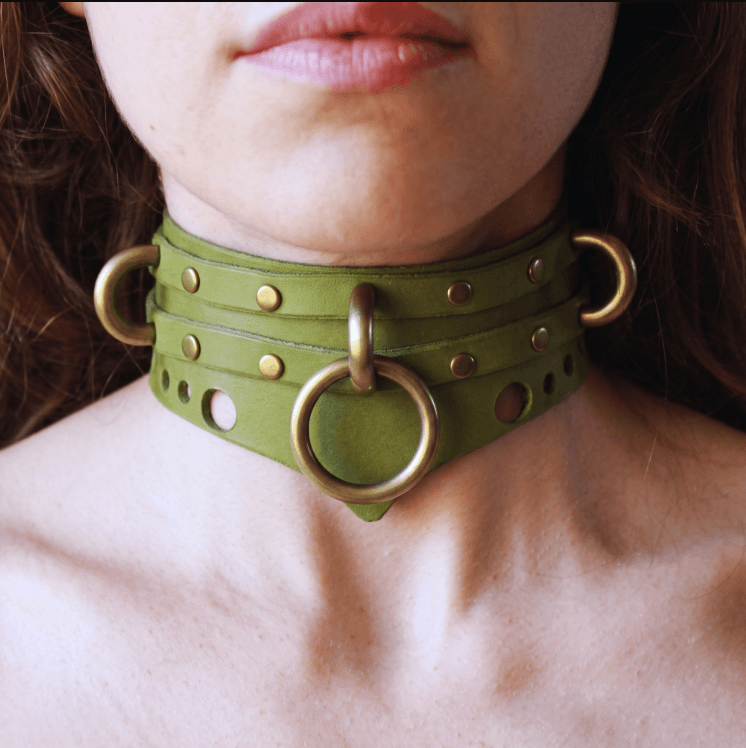 A close-up of a person's neck wearing a green leather choker necklace with metal rings and a metal ring.