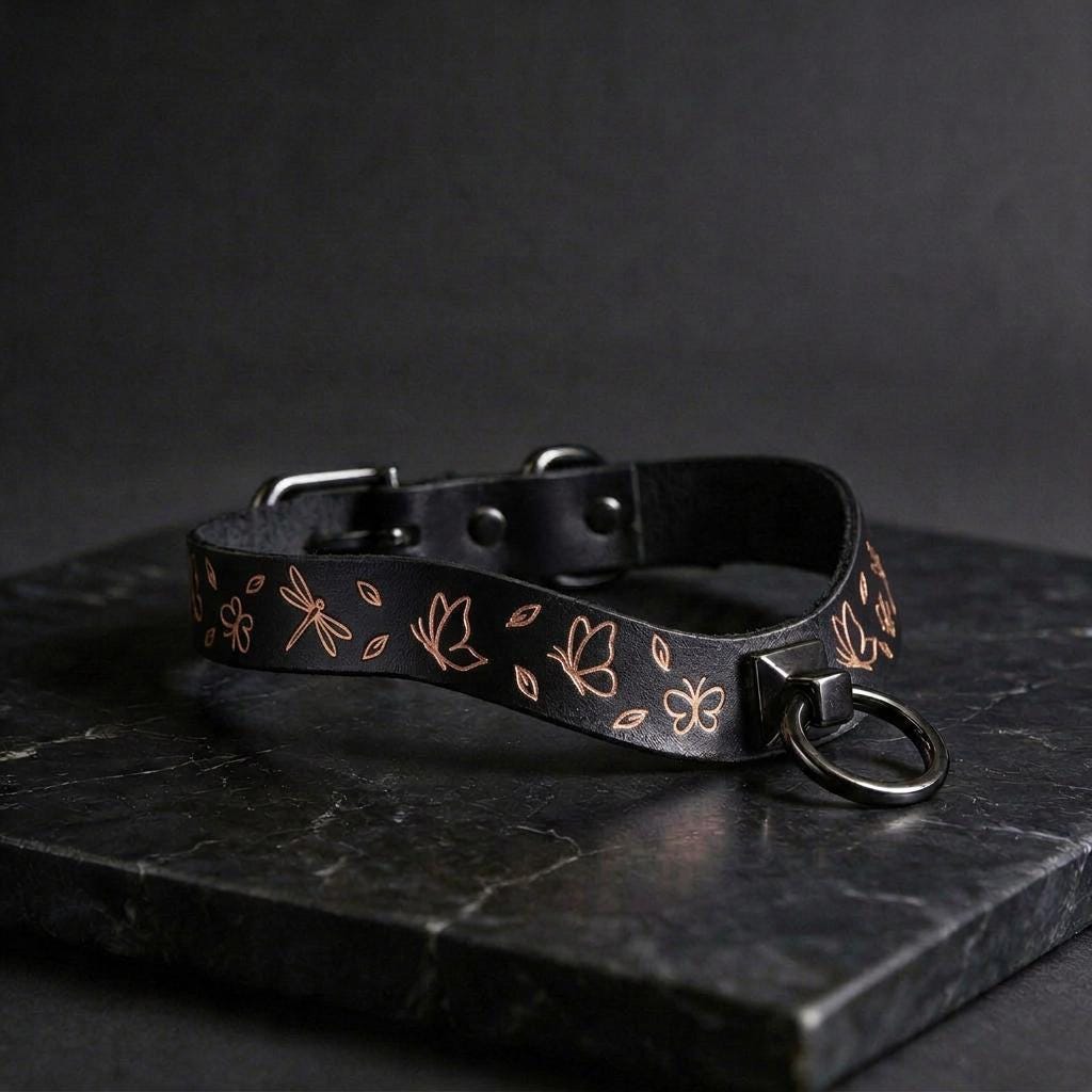 A black leather collar with a floral pattern and a metal ring on the front, resting on a dark surface.