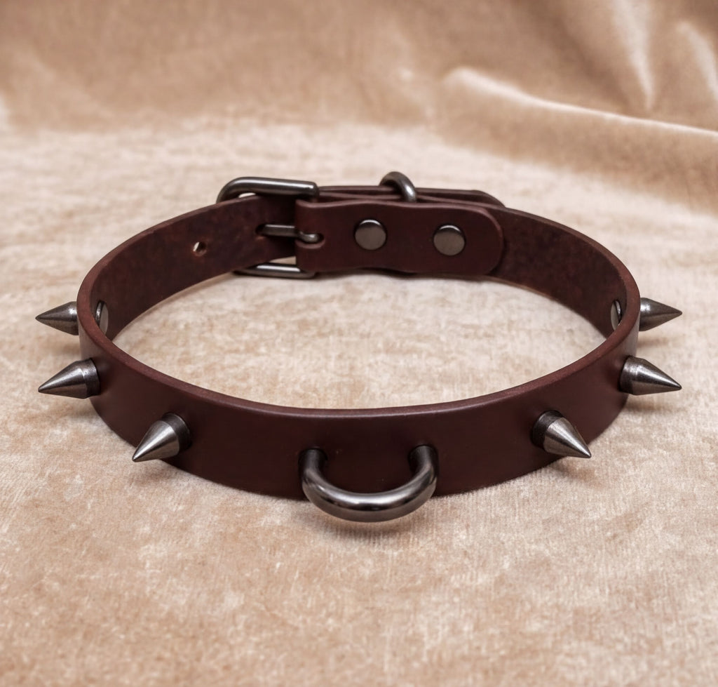 A brown leather spiked collar with metal spikes protruding from it, resting on a beige fabric background.