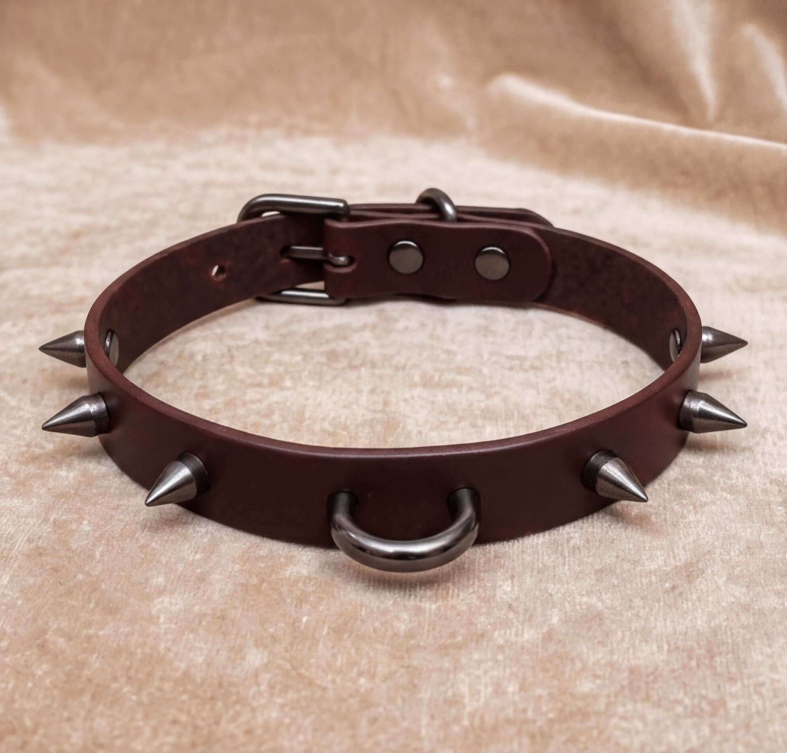 A brown leather spiked collar with metal spikes protruding from it, resting on a beige fabric background.