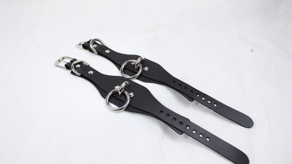 A pair of black leather straps with metal rings and buckles, lying on a white surface.