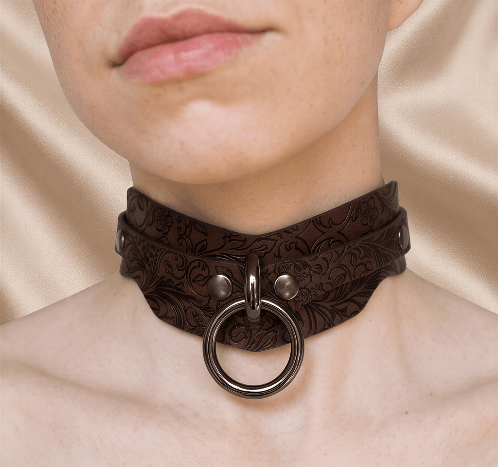 A close-up of a person's neck and chest area wearing a brown leather choker necklace with a metal ring.