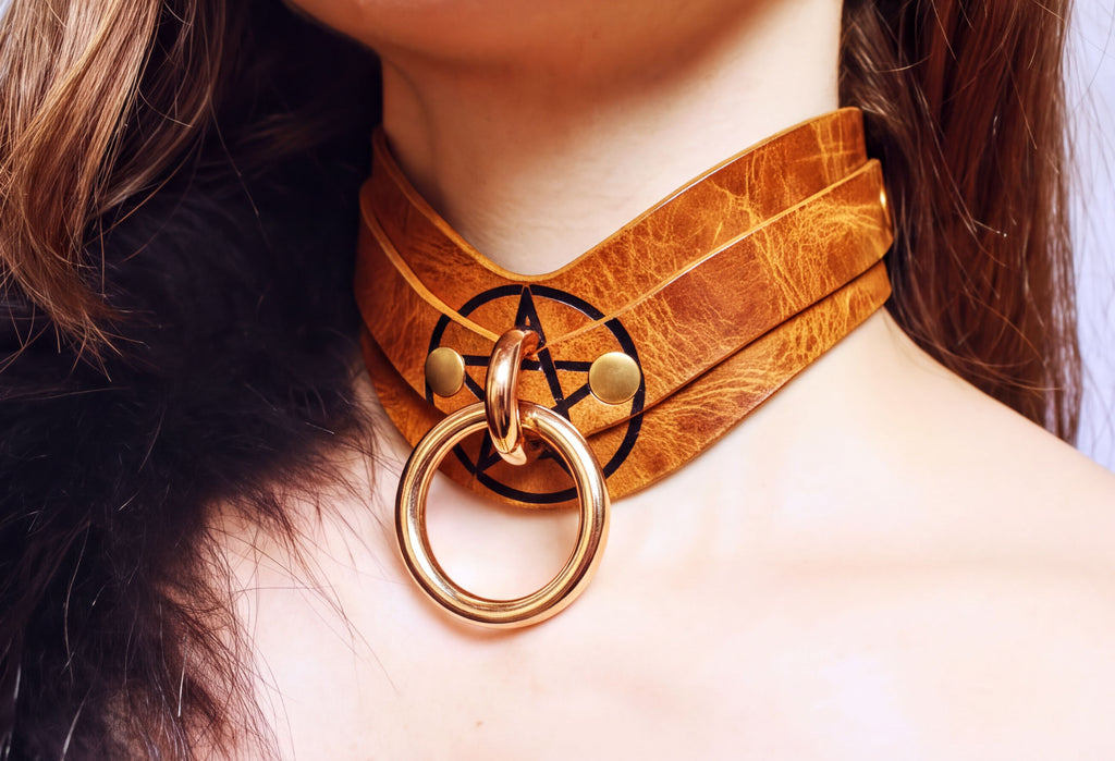 A close-up view of a woman's neck and chest area, featuring a choker-style leather collar with a gold ring and a symbol on it.