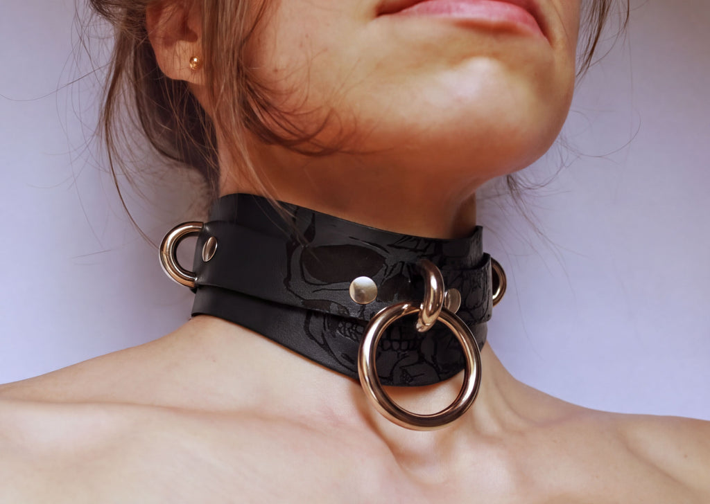 A close-up of a woman's neck and chest area wearing a black leather choker necklace with metal rings and a skull design.