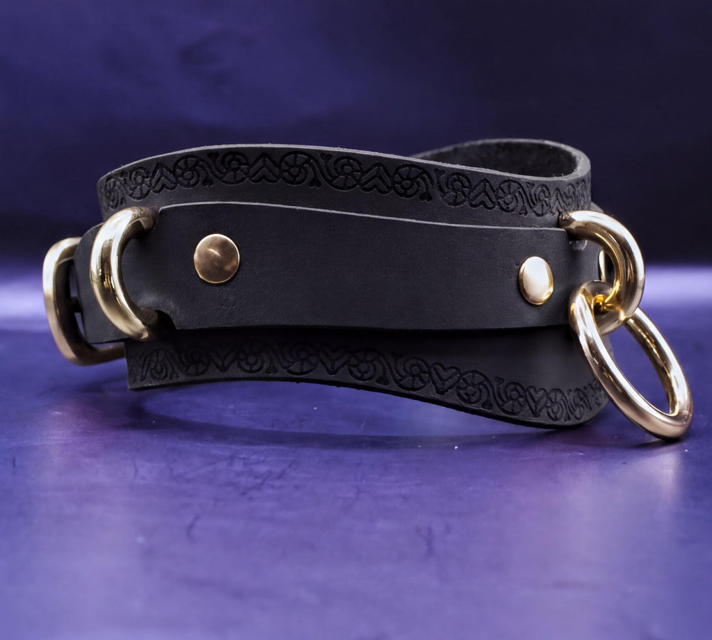 A black leather choker with gold metal rings and a decorative pattern on the edges, displayed on a purple background.