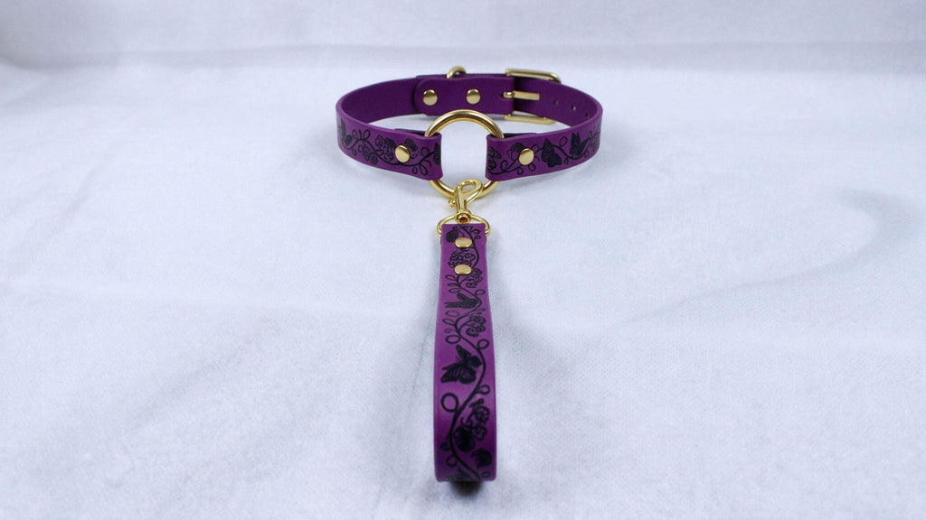 A purple collar with a gold buckle and a black floral pattern on the strap is displayed against a white background.