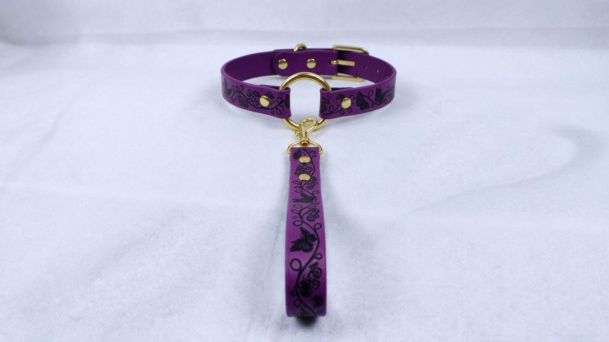 A purple collar with a gold buckle and a black floral pattern on the strap is displayed against a white background.