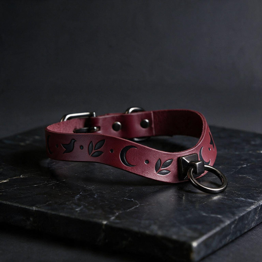 A burgundy leather collar with a moon and leaf design, featuring a metal ring and buckle, is displayed on a dark surface.
