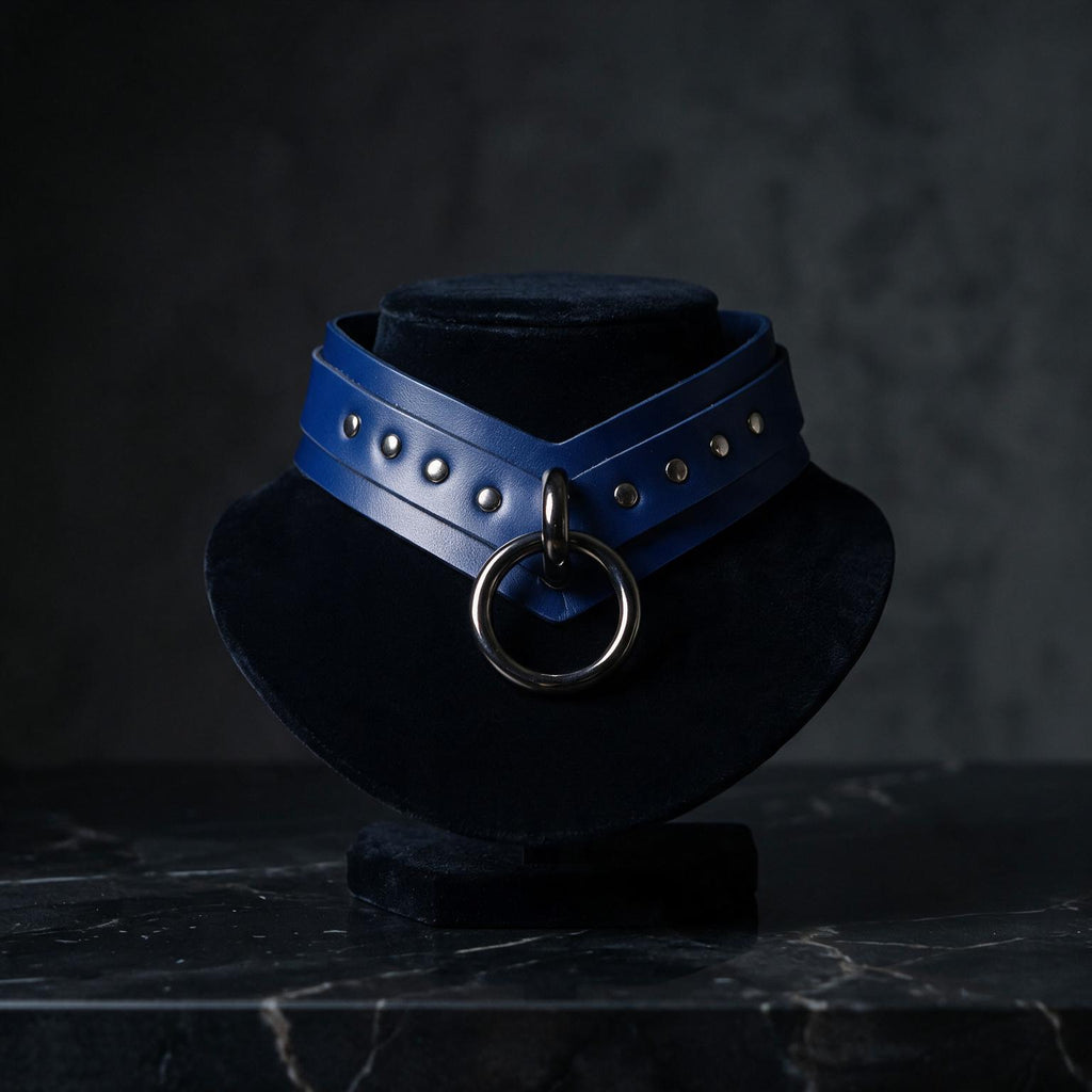 A blue leather choker necklace with a metal ring and studs is displayed on a black pedestal against a dark background.