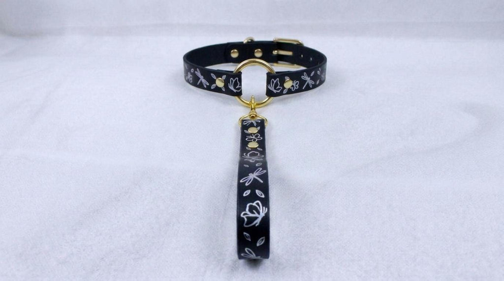 A black collar with a gold-colored buckle and a floral pattern on the strap is displayed against a white background.