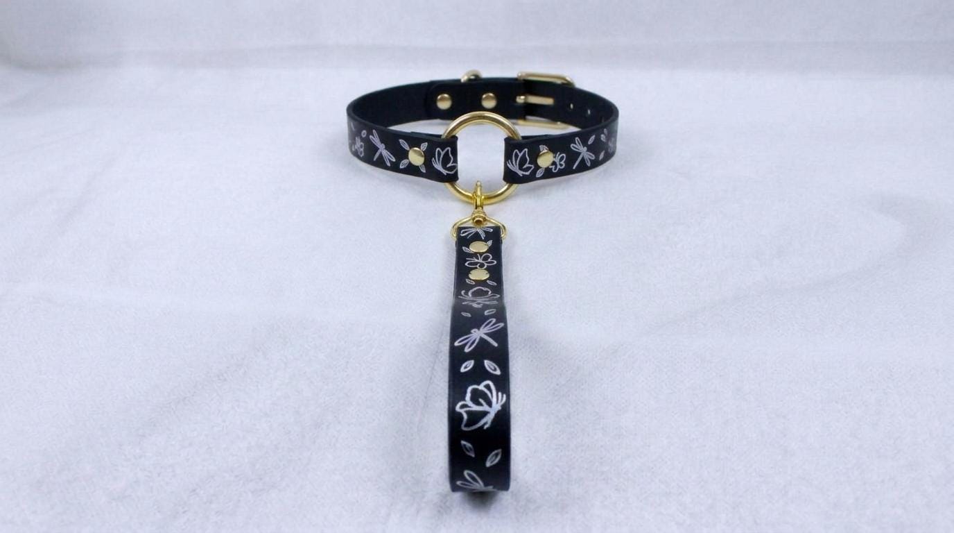 A black collar with a gold-colored buckle and a floral pattern on the strap is displayed against a white background.
