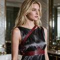 A woman with long blonde hair wearing a black dress with a red leather strap across her chest, standing in a room with a marble wall and a mirror.