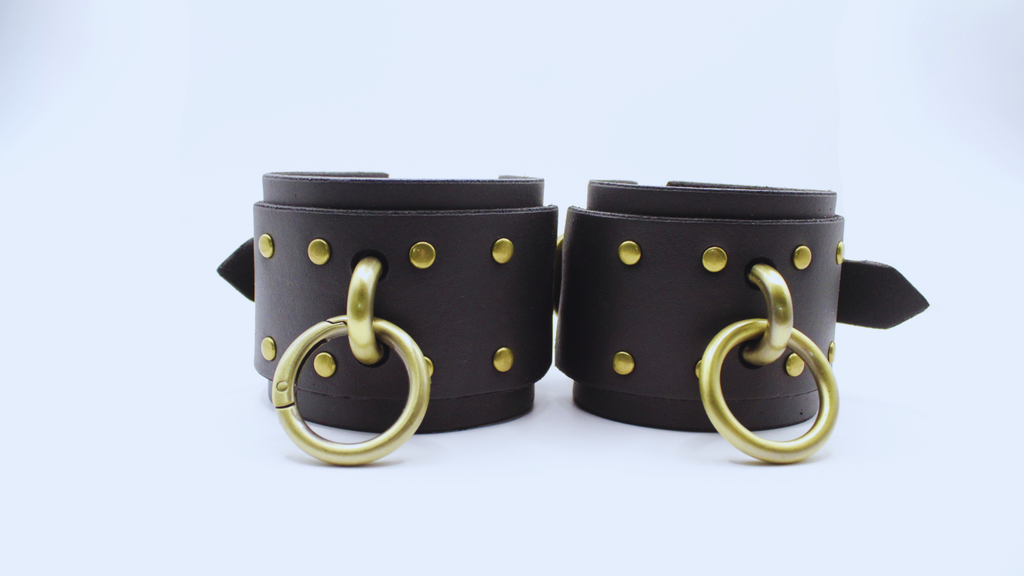 A pair of black leather cuffs with gold-colored metal rings and studs, set against a white background.