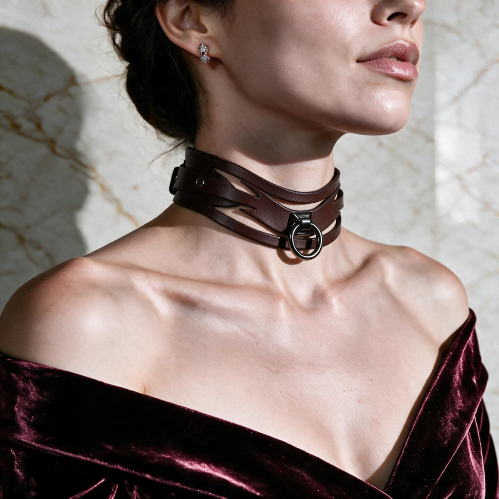 A woman wearing a brown leather choker necklace and a burgundy velvet off-the-shoulder dress stands against a marble wall.