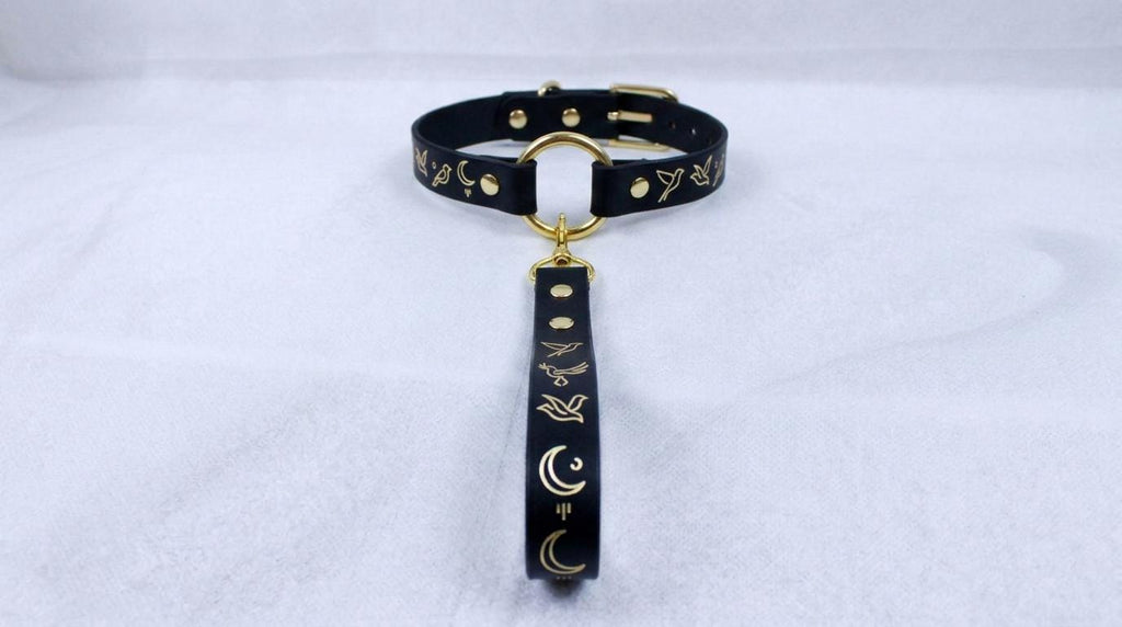 A black leather collar with gold accents and a gold-colored loop, featuring a design of birds and crescent moons.
