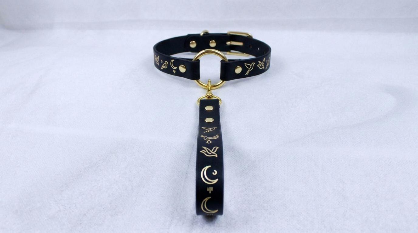 A black leather collar with gold accents and a gold-colored loop, featuring a design of birds and crescent moons.