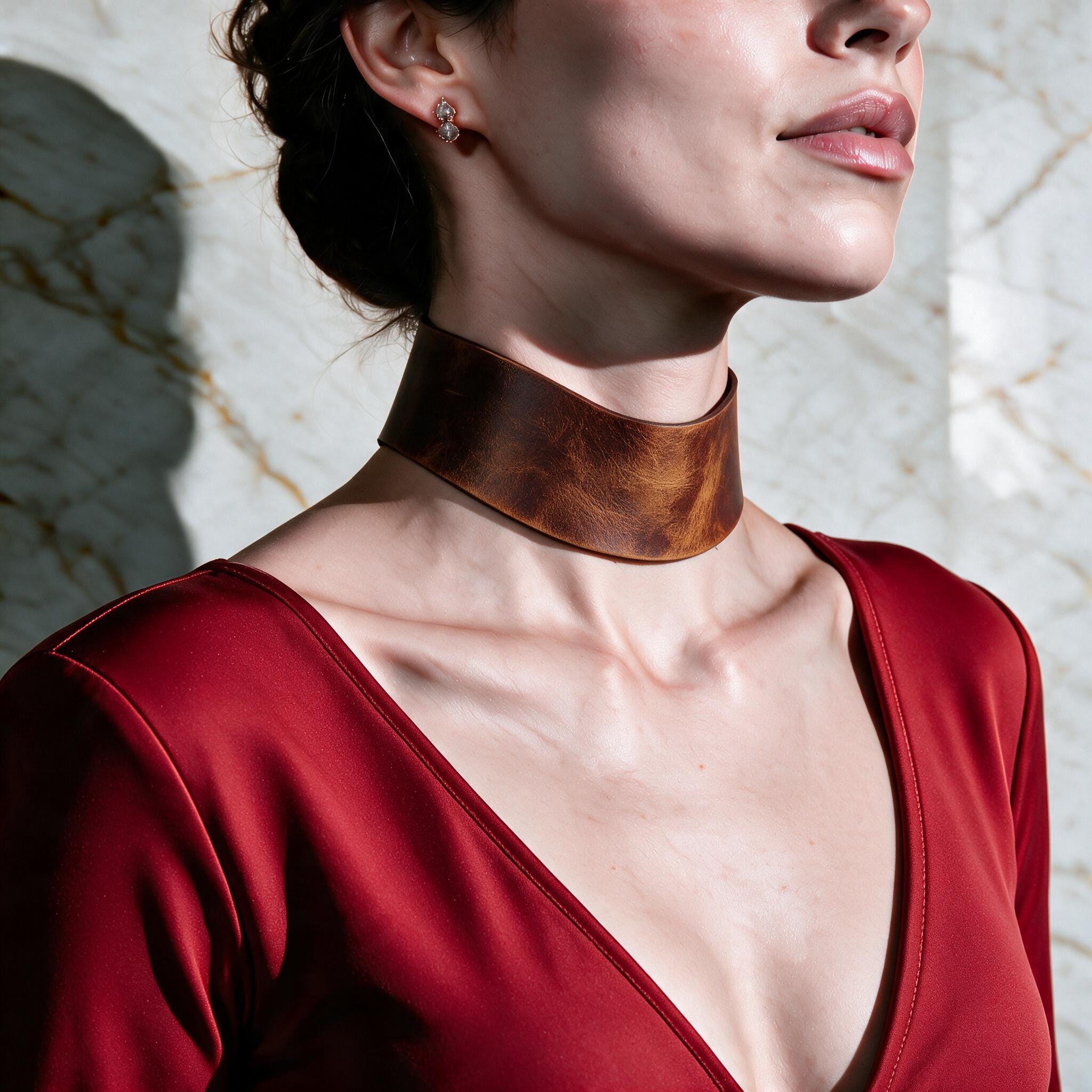 A woman wearing a red dress with a brown leather choker necklace stands against a marble wall.