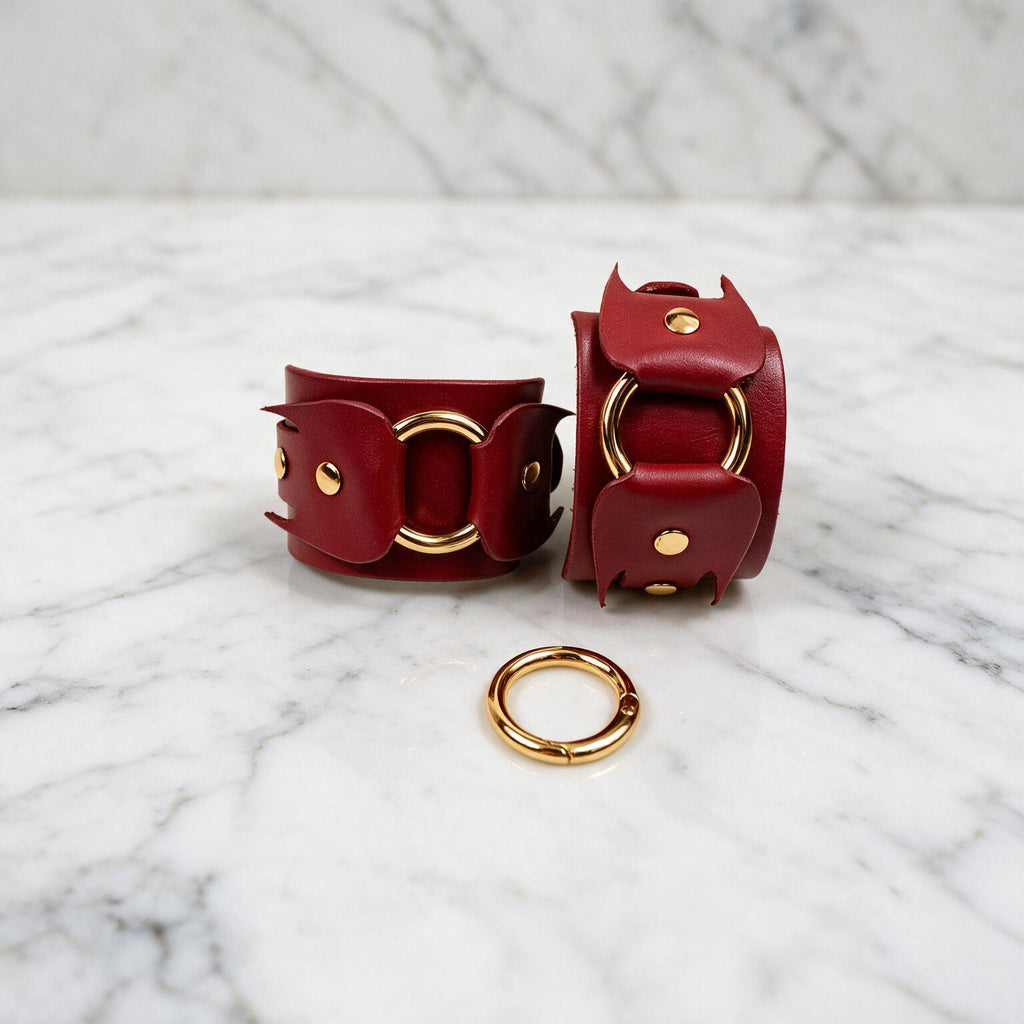 a pair of red leather cuffs with gold hardware, including a ring and a buckle, placed on a marble surface.
