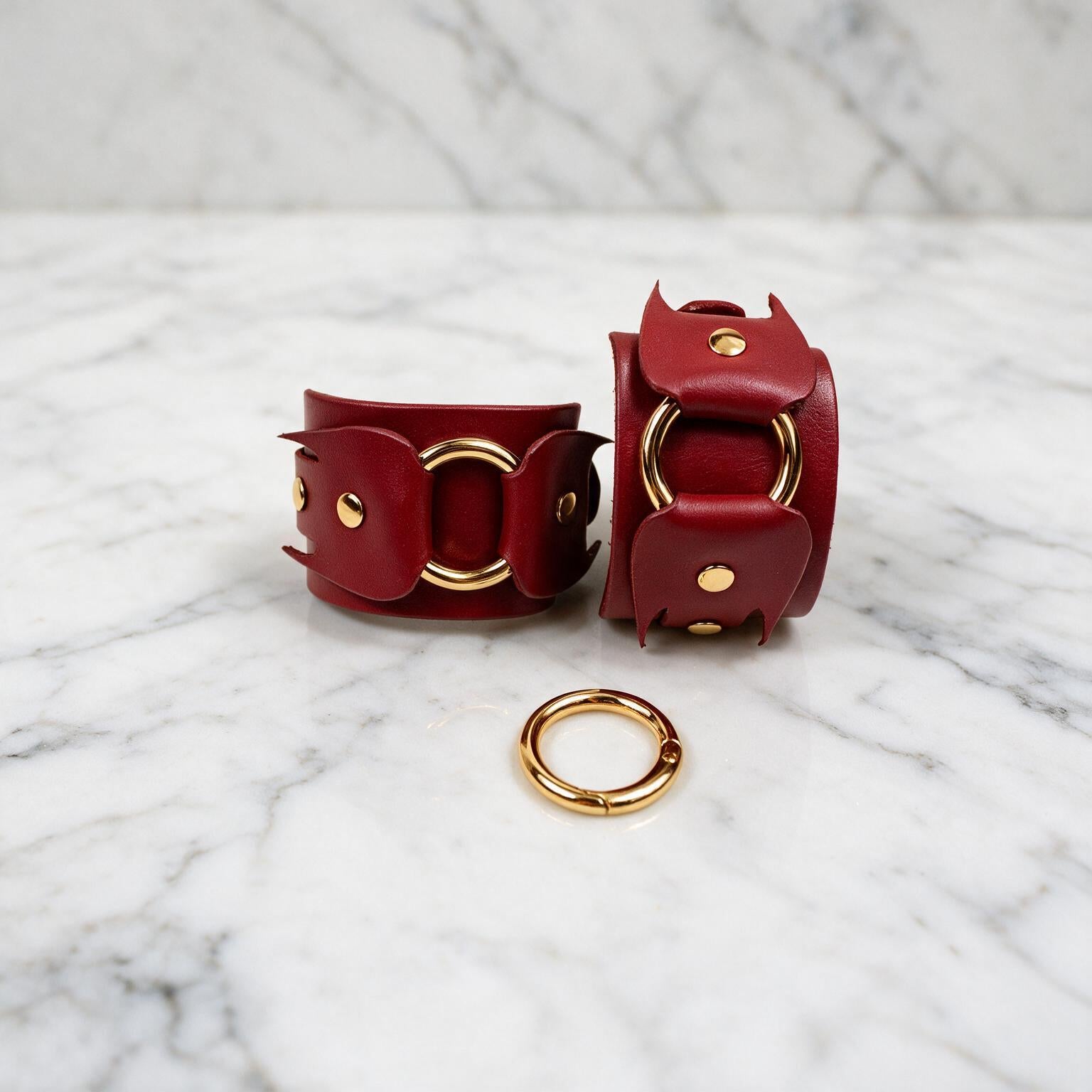 a pair of red leather cuffs with gold hardware, including a ring and a buckle, placed on a marble surface.