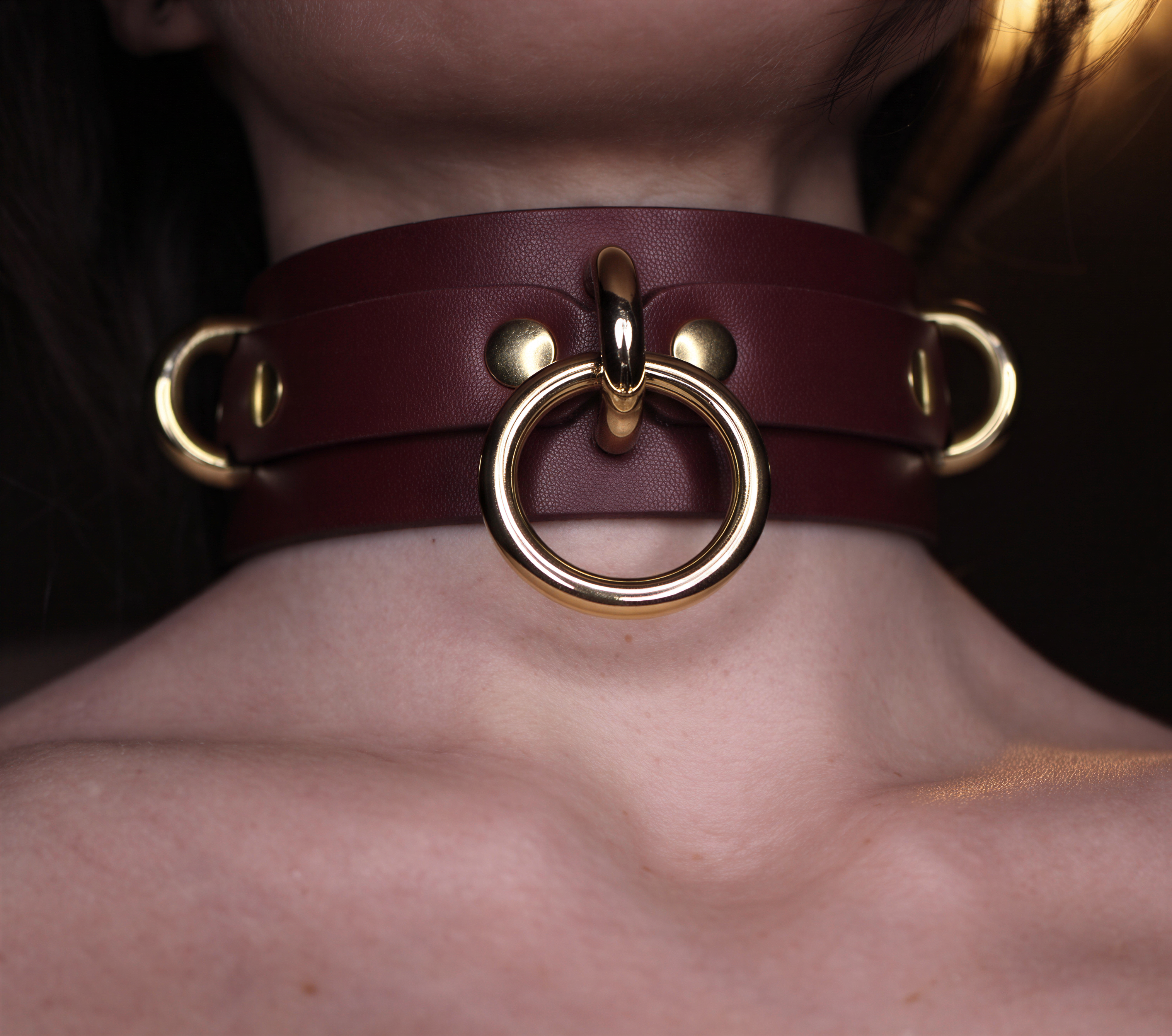 A close-up of a person's neck wearing a burgundy leather choker necklace with gold-colored metal rings.