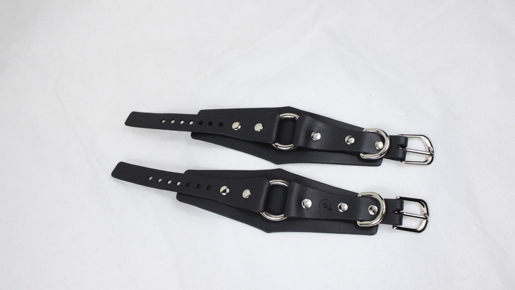 A pair of black leather straps with metal buckles and holes, laid out on a white surface.